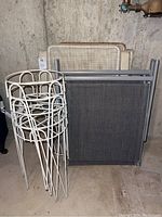 The lot has three folding chairs folded and standing upright, and four white metal plant stands stacked inside each other. The chairs include a lounger type and two Rio branded chairs with fabric and pockets on the back.