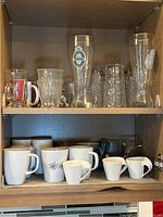 Top shelf with Crate & Barrel and other drinking glasses. Bottom shelf with white Villeroy & Boch espresso cups and IKEA mugs with floral designs.