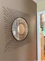 Full front view of the gold tone sunburst style wall mirror hung on a beige wall, showing its circular center mirror surrounded by radial metallic rods.