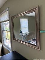 Mirror hung on a light-colored wall reflecting a window with blinds and an ironing board.
