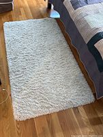 White shag area rug on hardwood floor beside a bed showing overall size and thickness.