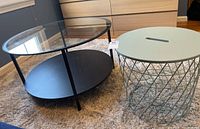 Two side tables on a carpeted floor. One is a black metal frame table with clear glass top and solid black bottom shelf. The other is a round light green table with a wire basket-style metal base and a solid top with a slit handle.