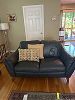 Front view of black leather sofa with two throw pillows (one beige, one patterned). Hardwood floor and interior home background.