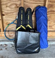 Photo showing two Armor brand badminton racket cases, a Head tennis racket, and a blue carrying bag in a garage setting.