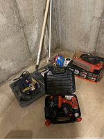 Photo shows a collection of power tools and cases including a Ryobi orbital jigsaw, Black & Decker cordless drill with charger and battery in a case, assorted drill bits and sandpaper tools with a wooden stick in the background against a concrete wall