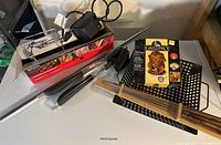 Photo showing BBQ set components on top of a white appliance, including universal rotisserie with motor and skewer, boxed chick toaster, black grill basket, black BBQ fork, black grill scraper, and four wooden-handled metal skewers.