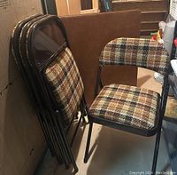 Four retro-style folding chairs with plaid upholstery in brown, gold, black, and cream colors, stacked against the wall in basement area.