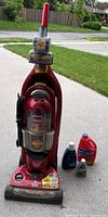 Full view of Bissell upright carpet cleaner with attached hose and bundled power cord, placed outside on a driveway. Three bottles of cleaning solutions are placed next to it.