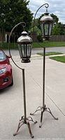Two tall metal hanging lanterns on curved metal poles with tripod bases outdoors. One lantern is taller than the other.