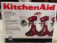 Box showing front view of KitchenAid Stand Mixer Attachment Pack with images of grinder and slicer attachments attached to red mixer, branding, and language variants.