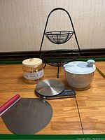 Two-tier black metal fruit stand with round baskets, Starfrit scale, white salad spinner, Cake Boss cake lifter, and glass cookie jar with plastic lid displayed on wood floor.