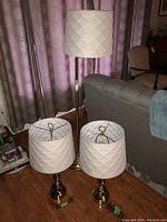 Photo showing one brass floor lamp with white textured drum shade and two matching brass table lamps with the same style shades, set on wooden floor near gray sofa.