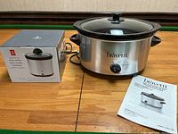 Bravetti 4L slow cooker placed on wooden surface with accompanying manual and box for PC mini slow cooker visible beside it.