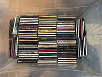 Photo of assorted music CDs stacked in a plastic bin, showing a variety of artist names and album titles on CD spines.