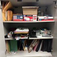 Full view of office supply cabinet filled with envelopes, folders, staplers, hole punches, tape holders, and calculator