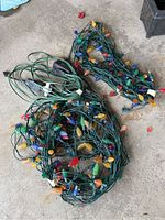 Two tangled strings of multi-colored outdoor Christmas light bulbs with green wiring, showing bulb variety and plugs.