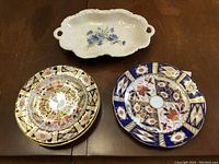 Photo of three plates: two Royal Crown Derby plates with detailed floral and geometric patterns, and one white ceramic floral tray with handles on wooden table.