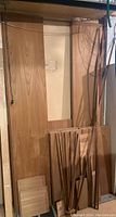 Photo showing a variety of wood pieces stacked against a beige wall in basement, including large panels and smaller strips mostly teak veneer particle board and some solid teak pieces.