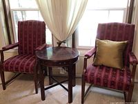 Two burgundy upholstered armchairs with patterned fabric and wooden frames, placed on either side of a round wooden Lane side table with minor scratches on top.