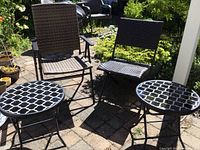 Two folding chairs with woven wicker-like seats and backs alongside two small round folding tables with black and white geometric glass tops on a patio.