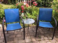 Two blue outdoor chairs with metal frames and small round glass-top table on brick patio with garden in background