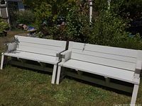 Two identical white plastic benches on grass in front of garden