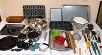 Wide shot showing the full assortment of baking dishes, pans, utensils and bowls laid out on a white sheet.