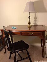 Full view of wooden desk with two drawers, brass lamp, and Tiffany pen on top, next to black vintage chair.