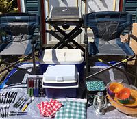 Overview of camping supplies including two Timber Ridge folding chairs, Cuisinart propane grill, two coolers, assorted cutlery, lighting, napkins, and plastic kitchenware.