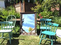 Overview photo showing four different blue wooden chairs and carved mirror outdoors on grass with trees and garden in background