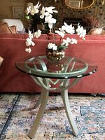 Front view of the round glass side table with white orchid floral arrangement on top, showing the metal base and thickness of the glass.