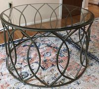 Clear beveled round glass top supported by metal decorative base with intertwined oval patterns, table shown on a patterned rug.