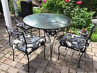 Full shot of round iron patio table with glass top and four cushioned chairs on brick patio surrounded by garden.