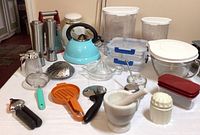Photo showing entire lot of kitchen items including kettle, storage containers, mortar and pestle, grinders, juicers, and various utensils on a white surface