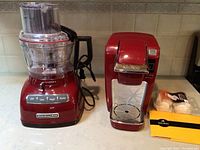 Red KitchenAid food processor and matching red Keurig coffee maker next to each other on a kitchen counter.