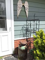 Photo showing white metal angel wings mounted on wall, antique milk can and black iron plant stand beside a door and by a bush