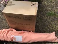 Cardboard box for 3-piece wicker conversation set with blue cushions (2 chairs and 1 table) seen next to coral umbrella in fabric bag.