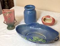 Four Roseville ceramic pieces displayed on white table: blue oblong bowl, blue vase, pink floral vase, reddish jar