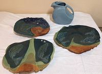 Three stoneware fish plates in dark blue, sage, and brown with powder blue Fiesta pitcher in background.