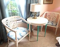 Two white painted wood armchairs with blue floral tapestry seats next to small round table with white top and turquoise legs. Vintage brass lamp on table. Part of seating area near window with curtains.