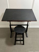 Black artist table with adjustable angled top and matching round stool beneath, photograph taken from front showing complete items.
