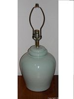 Photo of a heavy ceramic lamp in light blue with brass socket and harp, no bulb or shade, standing on floor against white wall.