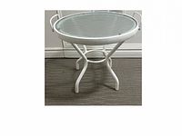 Round glass top table with white metal frame standing on carpeted floor against baseboard wall.