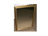 Image of a rectangular, gold-coloured framed mirror leaning against a wall on a beige carpet. The mirror surface is clear with no visible scratches or damage. The gold frame has ornate decorative inner border details.