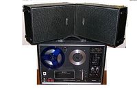 Front view of Sony TC-270 reel-to-reel tape deck with two attached speakers set upright behind it. The tape deck is black with a silver circular reel area and control panel to the right.
