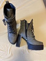 Pair of women’s heeled combat boots in greyish color with lace-up front, side zipper, thick platform sole, and block heel viewed from top and side