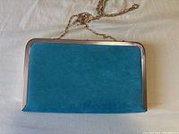 Front view of a rectangular blue suede clutch purse with a silver-tone metal frame and gold chain strap laid out on a white surface.