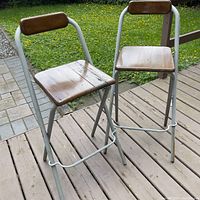 Two wooden and metal framed bar stools positioned outdoors on wood decking and concrete paving, showing full view.