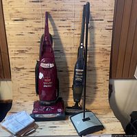 Photo showing two upright vacuum cleaners side by side, a maroon Bissell PowerGlide Platinum and a black Hoover model, with a manual floor sweeper and papers in front.