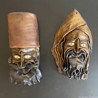 Front view of two handcrafted leather masks on black background, showing detailed carved facial features and headgear.